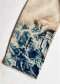 A close-up of a cream-colored fabric garment laid flat on a white background, showing a blue and white roman sun printed design concentrated on the lower portion.