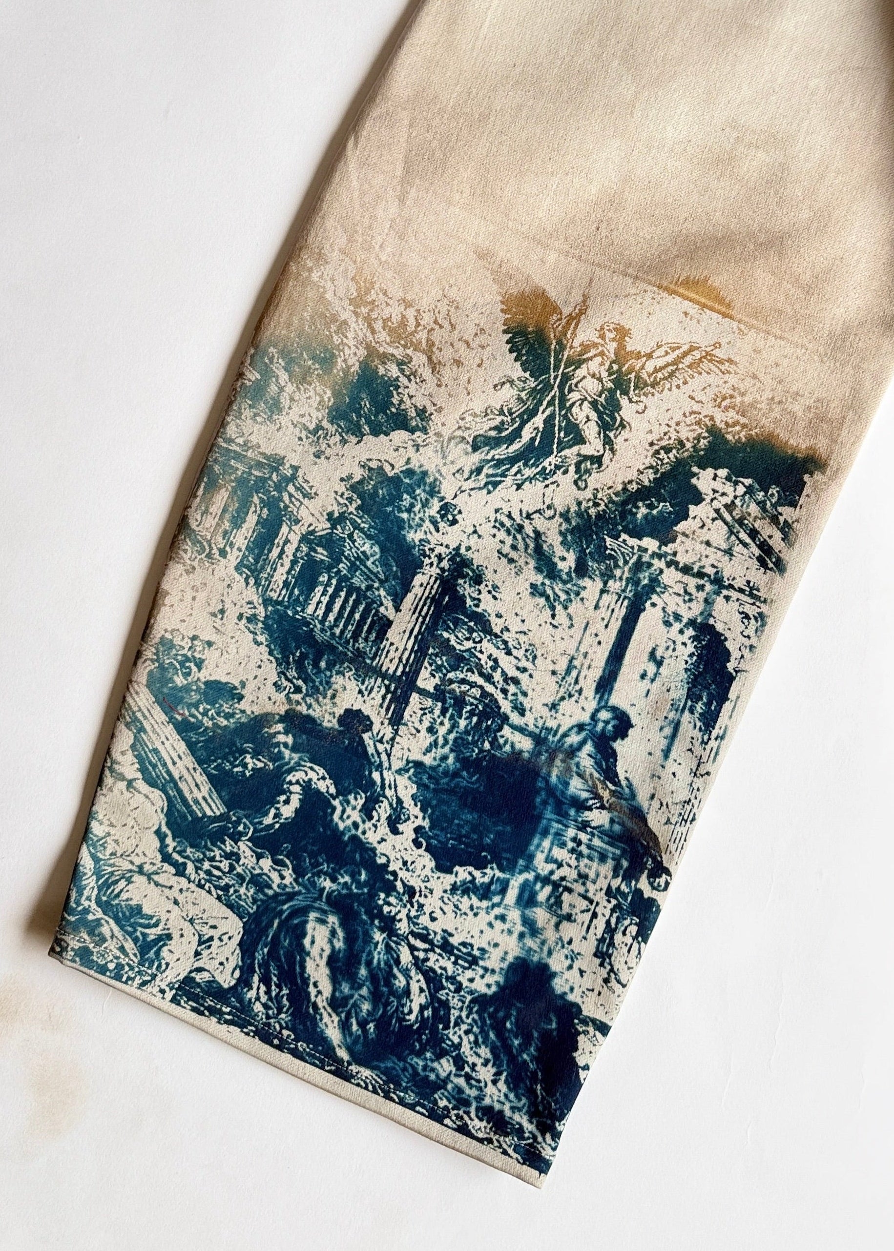 A close-up of a cream-colored fabric garment laid flat on a white background, showing a blue and white roman sun printed design concentrated on the lower portion.